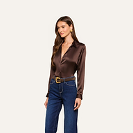 Wearing the Tyler Silk Blouse in dark brown paired with blue high-waisted jeans, a brown belt featuring a gold buckle, and gold earrings, a woman stands against an off-white background.