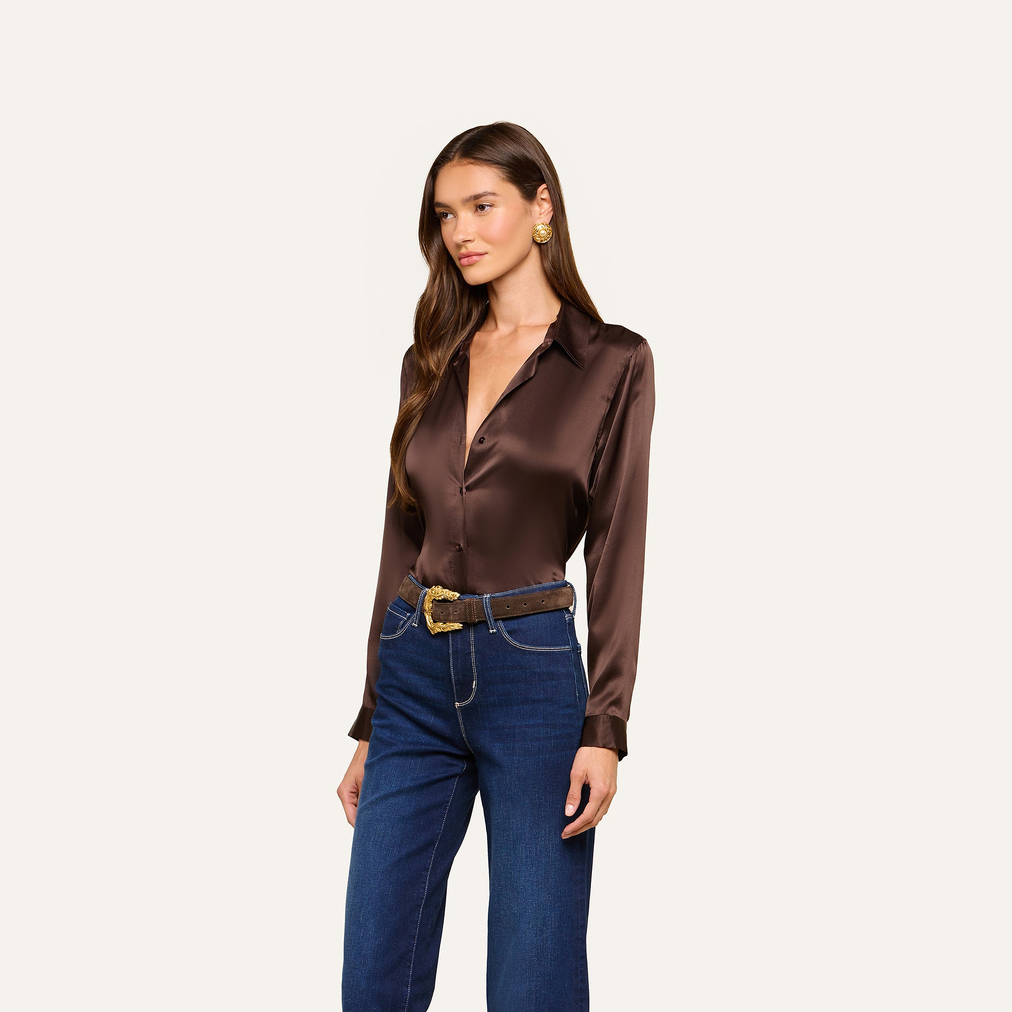 Wearing the Tyler Silk Blouse in dark brown paired with blue high-waisted jeans, a brown belt featuring a gold buckle, and gold earrings, a woman stands against an off-white background.
