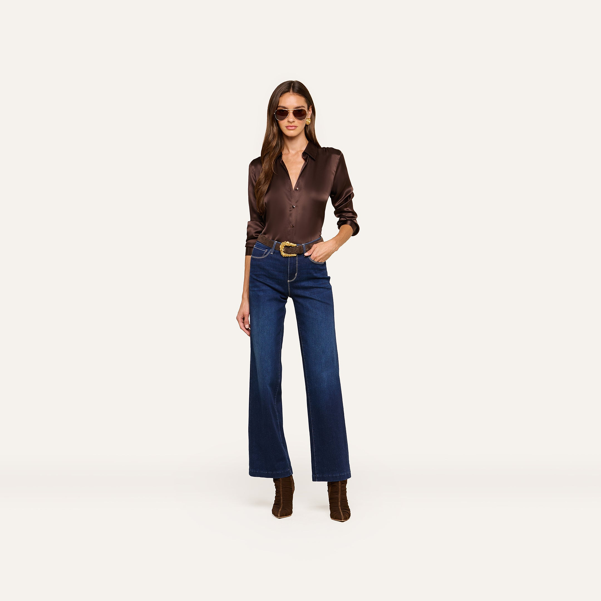 A woman wearing sunglasses, the Tyler Silk Blouse in dark brown charmeuse, blue high-waisted wide-leg jeans, a brown belt, and brown heeled boots stands against a plain white background.