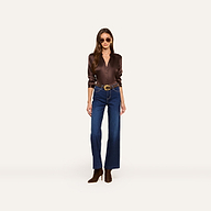 Wearing the Tyler Silk Blouse in a glossy brown hue, high-waisted blue jeans, brown boots, a wide belt, and sunglasses, a woman stands against a plain white background.