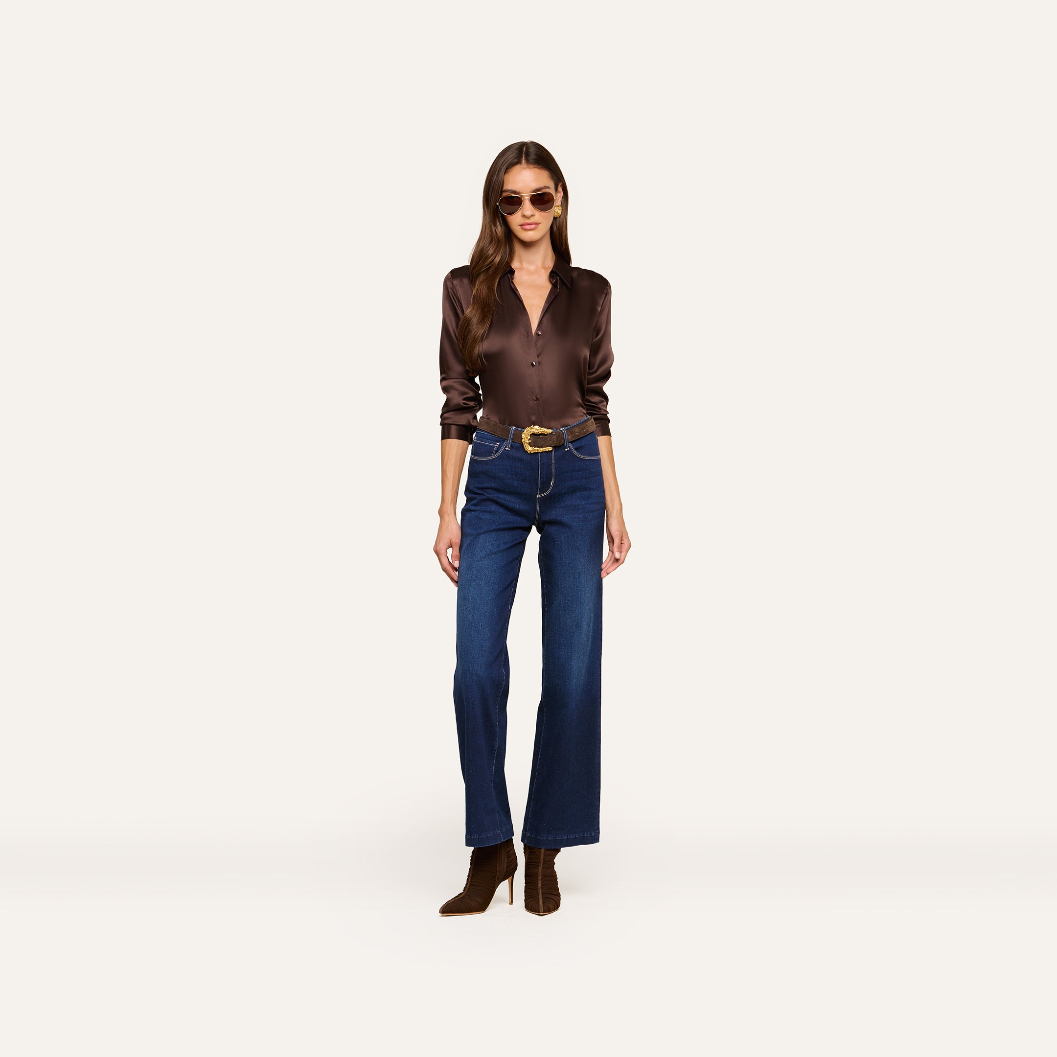 Wearing the Tyler Silk Blouse in a glossy brown hue, high-waisted blue jeans, brown boots, a wide belt, and sunglasses, a woman stands against a plain white background.