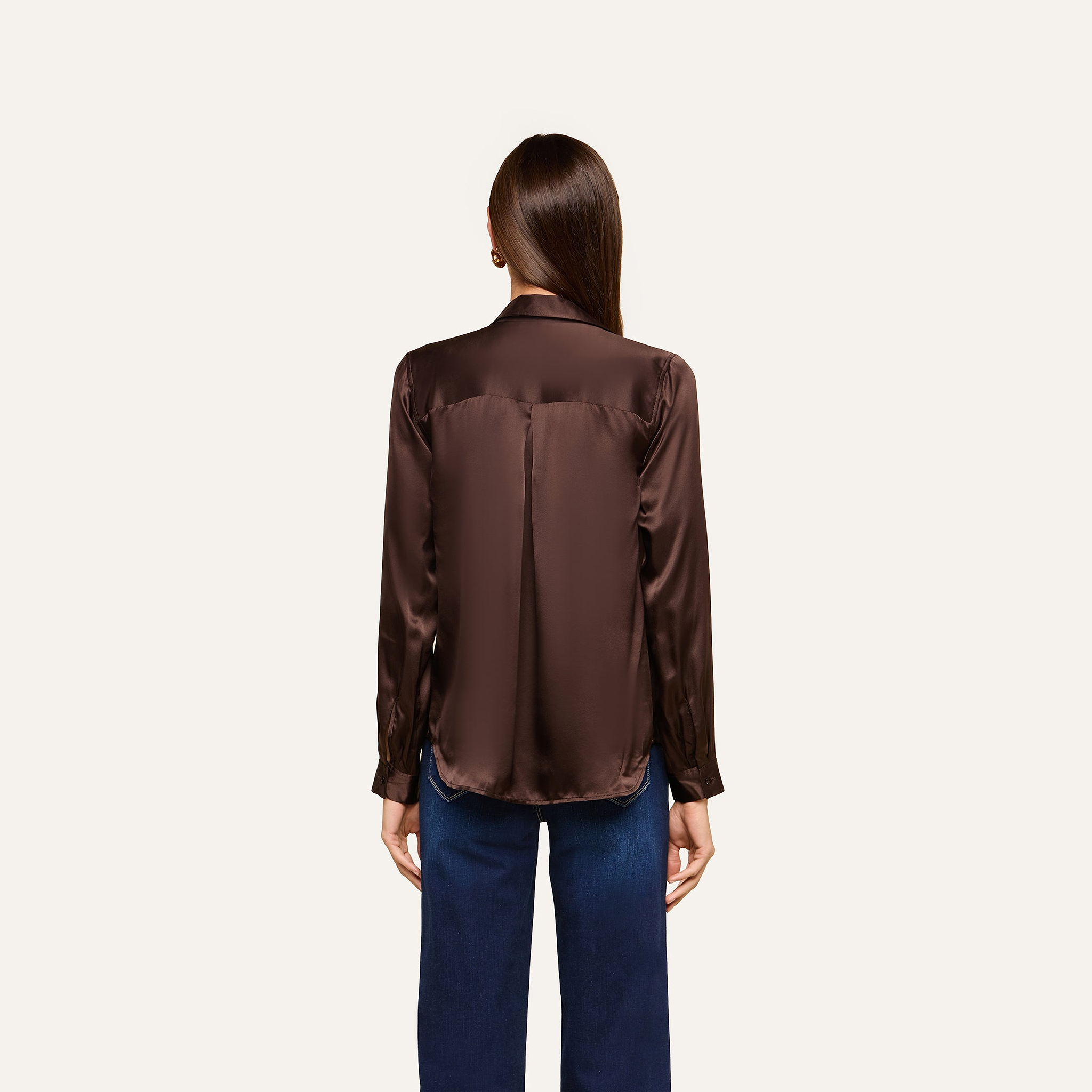 A woman with long brown hair, seen from the back, is wearing a dark brown Tyler Silk Blouse with long sleeves and blue pants, standing against a plain light background.