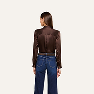 A woman with long brown hair, dressed in the Tyler Silk Blouse in dark brown and blue jeans, stands facing away from the camera against a plain light background.