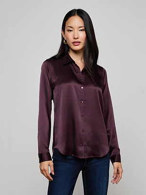 A woman with long dark hair wears the L'AGENCE Tyler Silk Blouse in deep purple with long sleeves, paired with dark blue jeans, standing against a plain light gray background.