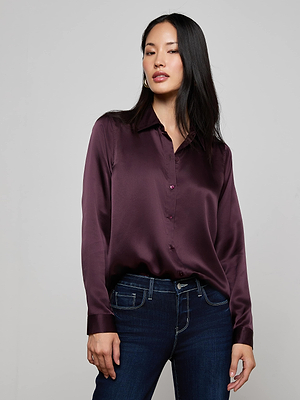A woman with long black hair wears the L'AGENCE Tyler Silk Blouse in dark purple and dark blue jeans, standing with one hand by her side against a plain light gray background.