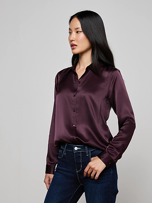A woman with long black hair wears the L'AGENCE Tyler Silk Blouse in dark purple and dark blue jeans, standing against a light gray background and looking to her left.