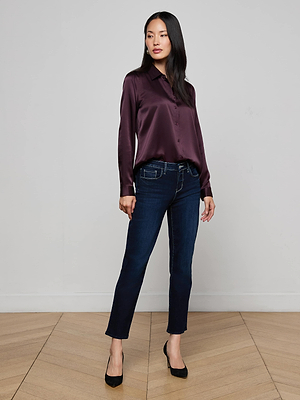 A woman stands in a modern room wearing the L’AGENCE Tyler Silk Blouse, styled with dark blue ankle jeans and black heels. She poses with one hand on her hip, looking slightly to the side.