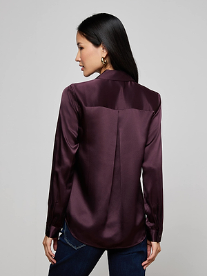 A woman with long dark hair and gold hoop earrings models the L'AGENCE Tyler Silk Blouse in dark purple with dark jeans. She faces away from the camera, displaying the back of the blouse.