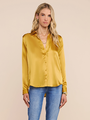 A woman with long blonde hair wears the shiny gold Tyler Silk Blouse and blue jeans, standing against a plain beige background.