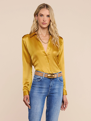 A woman with long blonde hair wears the shiny gold Tyler Silk Blouse by L’AGENCE, featuring long sleeves and button-down front, tucked into blue jeans with a tan belt. She stands against a plain beige background, looking at the camera.