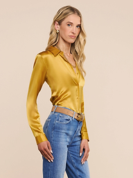 A woman with long blonde hair stands sideways against a plain light background, looking at the camera with a neutral expression. She is wearing the Tyler Silk Blouse—a long-sleeve, gold, button-down top—paired with blue jeans.