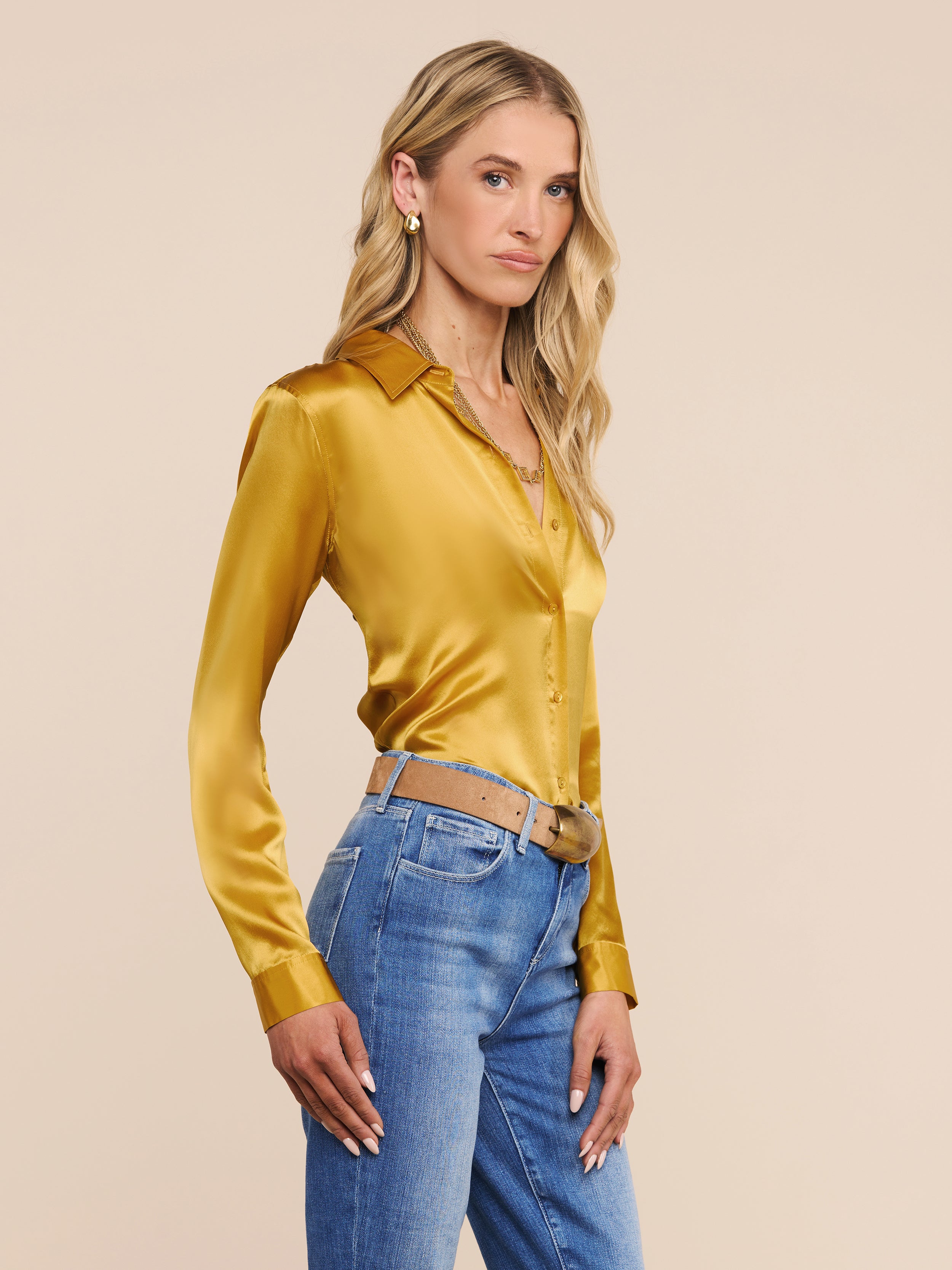 A woman with long blonde hair stands sideways against a plain light background, looking at the camera with a neutral expression. She is wearing the Tyler Silk Blouse—a long-sleeve, gold, button-down top—paired with blue jeans.
