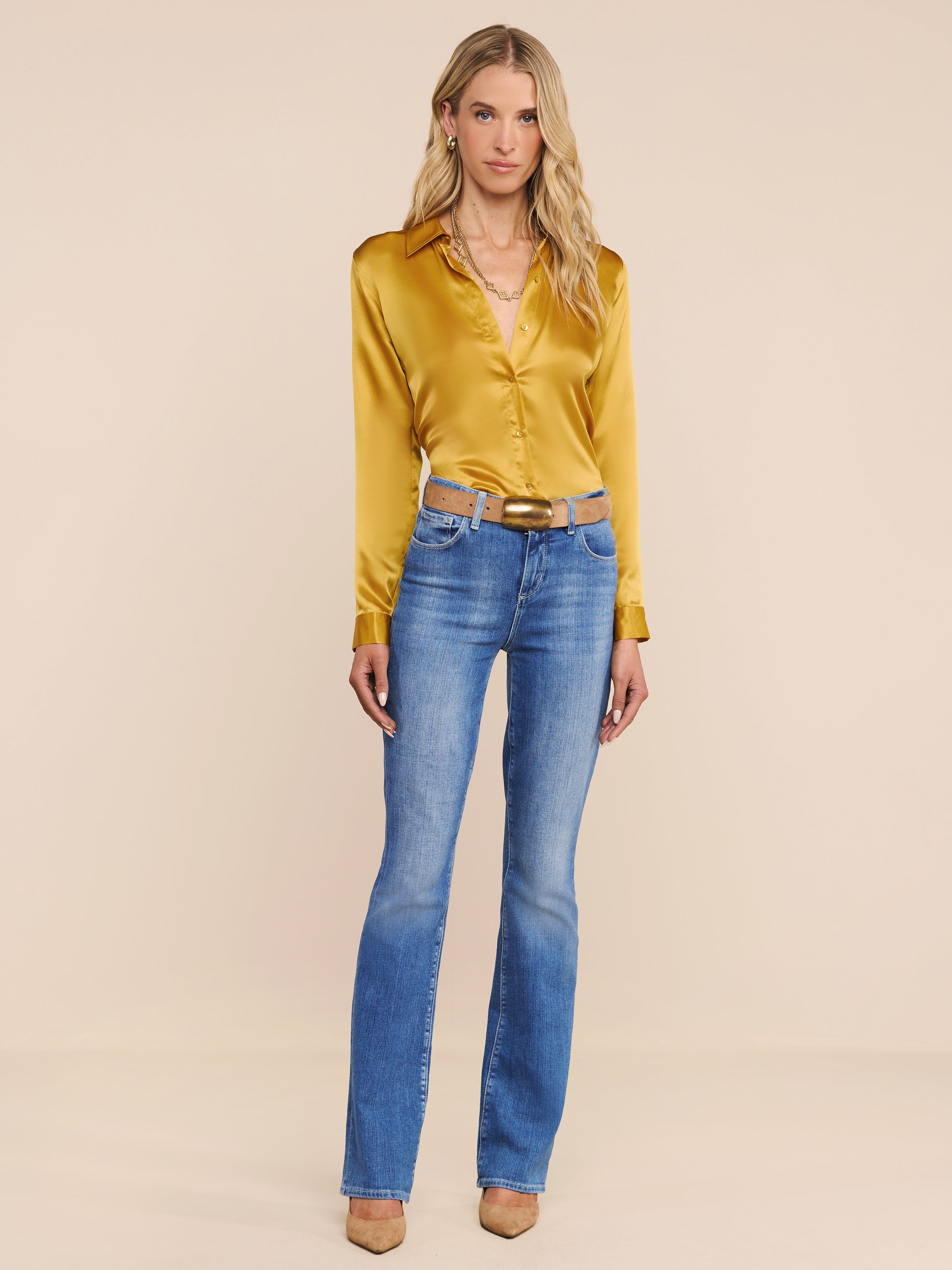 A woman stands against a plain white background wearing the shiny yellow Tyler Silk Blouse, blue jeans with a belt, and tan heels. She has long blonde hair and a neutral expression.