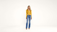 A woman stands in a bright, minimal space wearing a shiny yellow blouse, blue jeans, and tan heels. She has long blonde hair and a relaxed expression.