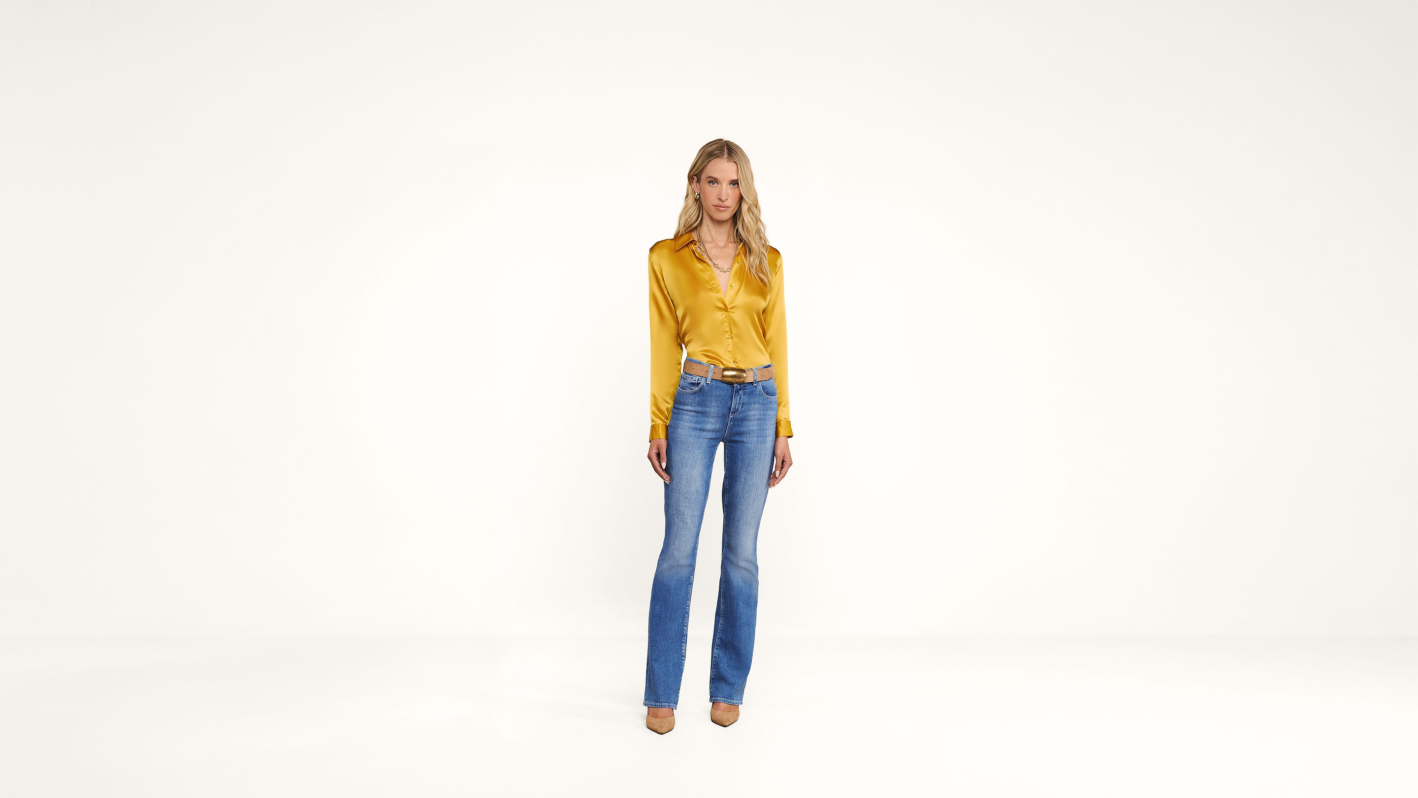 A woman stands in a bright, minimal space wearing a shiny yellow blouse, blue jeans, and tan heels. She has long blonde hair and a relaxed expression.
