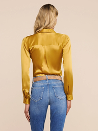 A woman stands facing away, wearing the shiny gold Tyler Silk Blouse from L’AGENCE paired with blue jeans featuring visible back pockets, set against a plain light background.