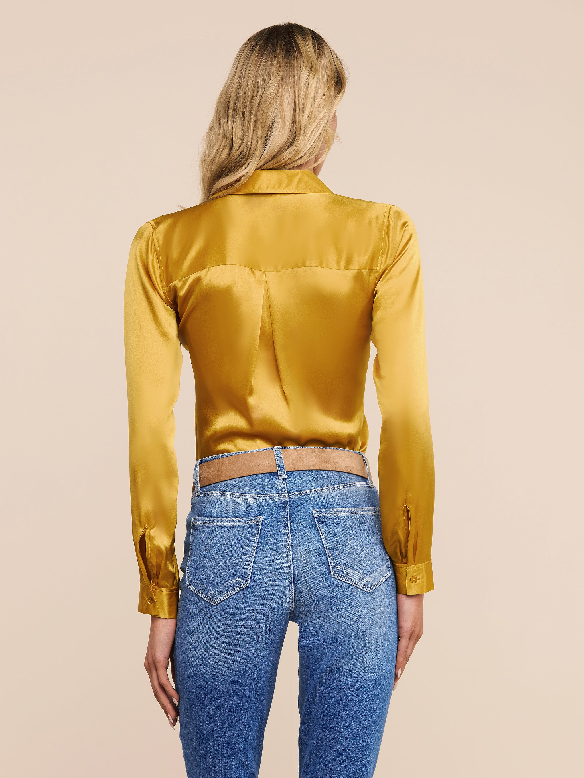 A woman stands facing away, wearing the shiny gold Tyler Silk Blouse from L’AGENCE paired with blue jeans featuring visible back pockets, set against a plain light background.