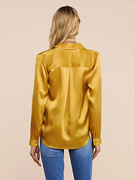 A woman with long blonde hair, seen from behind, is wearing a shiny golden yellow Tyler Silk Blouse paired with blue jeans, standing against a plain light background.
