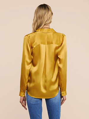 A woman with long blonde hair is shown from behind, wearing a shiny gold Tyler Silk Blouse and blue jeans against a plain light beige background.