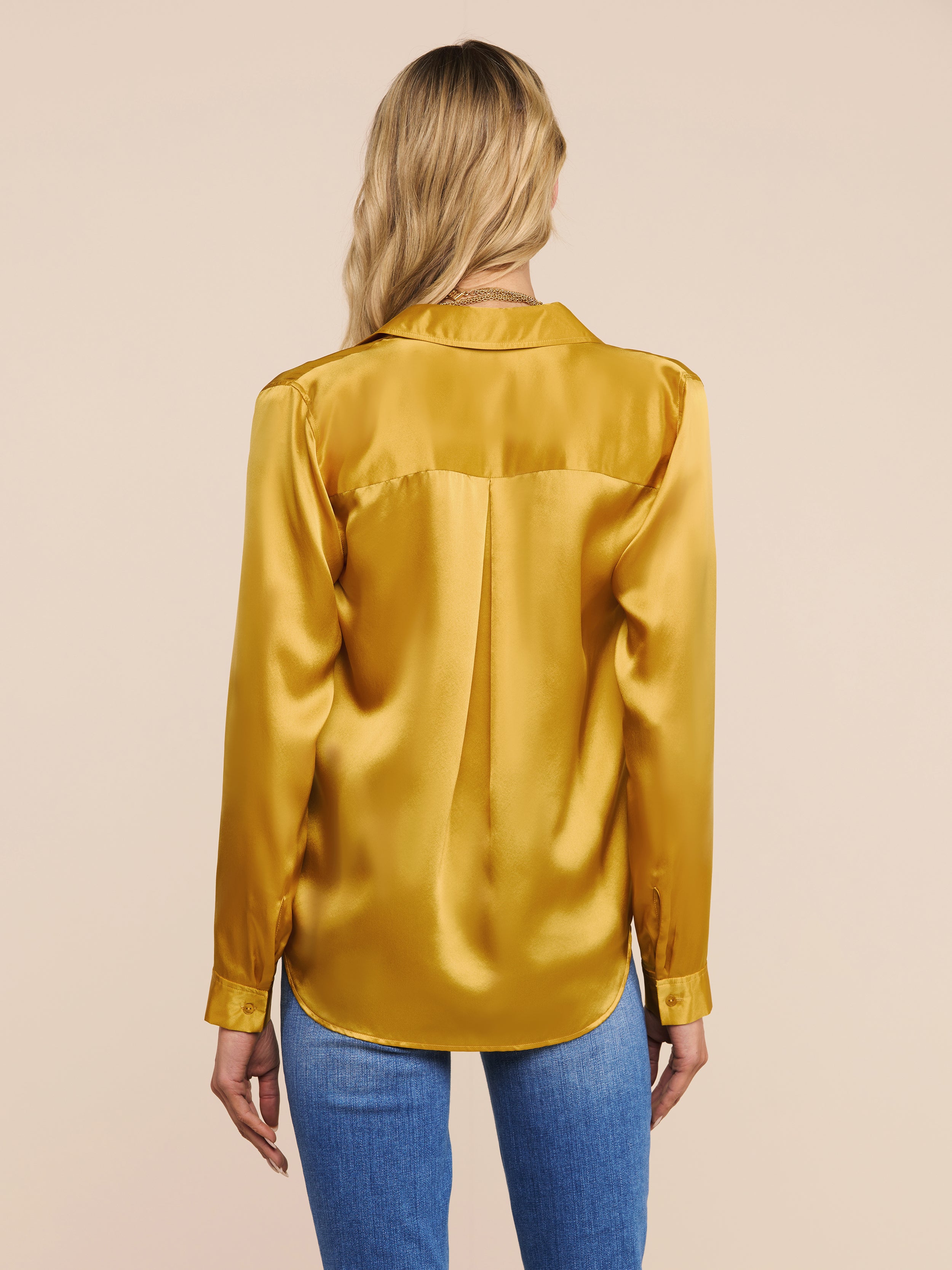 A woman with long blonde hair, seen from behind, is wearing a shiny golden yellow Tyler Silk Blouse paired with blue jeans, standing against a plain light background.