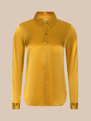 The Tyler Silk Blouse by LAGENCE is a long-sleeve, golden yellow silk button-down with a pointed collar and sleek finish, shown against a plain beige background.