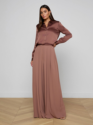 A woman stands indoors wearing the Tyler Silk Blouse and matching high-waisted, wide-leg pants in dusty rose, posing with one hand on her hip against a plain background.