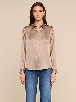 A woman with long brown hair stands facing forward against a plain beige background, wearing the shiny cardamom brown Tyler Silk Blouse and blue jeans.