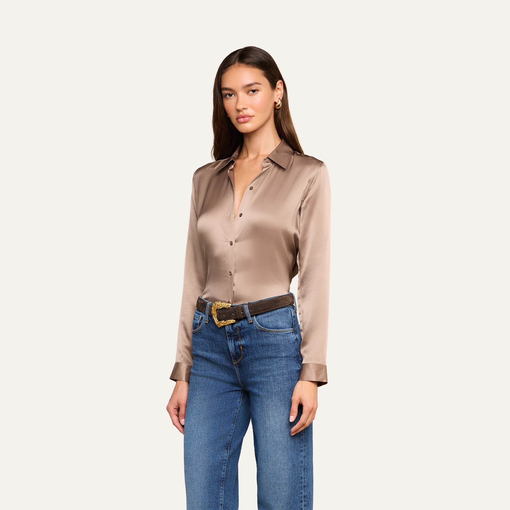 A woman wearing the Tyler Silk Blouse in cardamom brown, styled with blue jeans and a brown belt, stands against a plain light background.