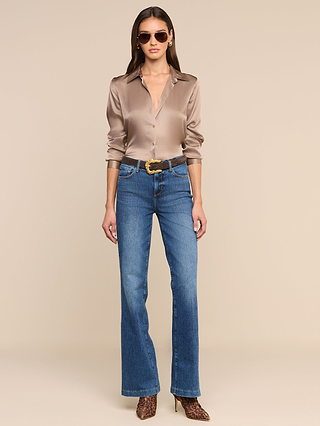 A woman stands facing forward against a beige background, wearing the Tyler Silk Blouse in shiny cardamom brown, styled with blue high-waisted jeans, a brown belt, leopard print heels, and sunglasses.