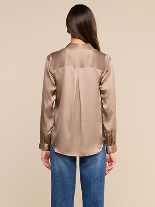 A person with long brown hair, dressed in the Tyler Silk Blouse in a shiny cardamom brown shade and blue jeans, is standing with their back to the camera against a plain beige background.