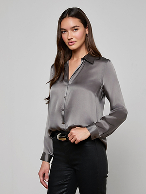 A woman with long brown hair wears the Tyler Silk Blouse in shiny gray and black pants, standing against a plain light gray background. She looks at the camera with a slight smile and relaxed pose.