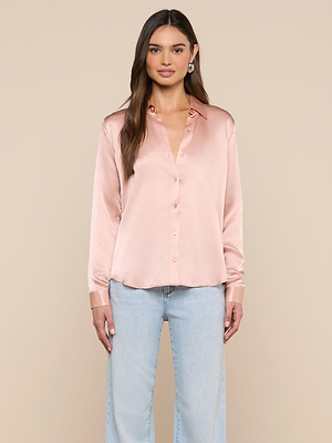 A woman faces forward in a shiny light pink Tyler Silk Blouse with long sleeves and light blue jeans, set against a solid beige background.
