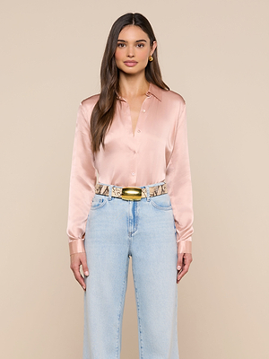 A woman with long brown hair wears a light pink Tyler Silk Blouse, light blue high-waisted jeans, a snakeskin belt with a gold buckle, and gold earrings, standing against a beige background.