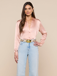 A woman with long brown hair wears the LAGENCE Tyler Silk Blouse in pale pink, tucked into high-waisted light blue jeans with a snakeskin-patterned belt and gold buckle, standing against a beige background.