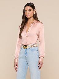 A woman with long brown hair stands against a beige background, wearing the L’AGENCE Tyler Silk Blouse in light pink, light blue high-waisted jeans, and a snakeskin-patterned belt with a gold buckle.