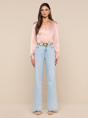 A woman with long brown hair stands against a beige background, wearing the LAGENCE Tyler Silk Blouse in light pink, light blue high-waisted wide-leg jeans, a beige snake print belt, and nude heels. She has a neutral expression.