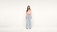 A woman stands facing forward against a plain white background, wearing a light pink blouse, light blue high-waisted wide-leg jeans, and beige shoes.