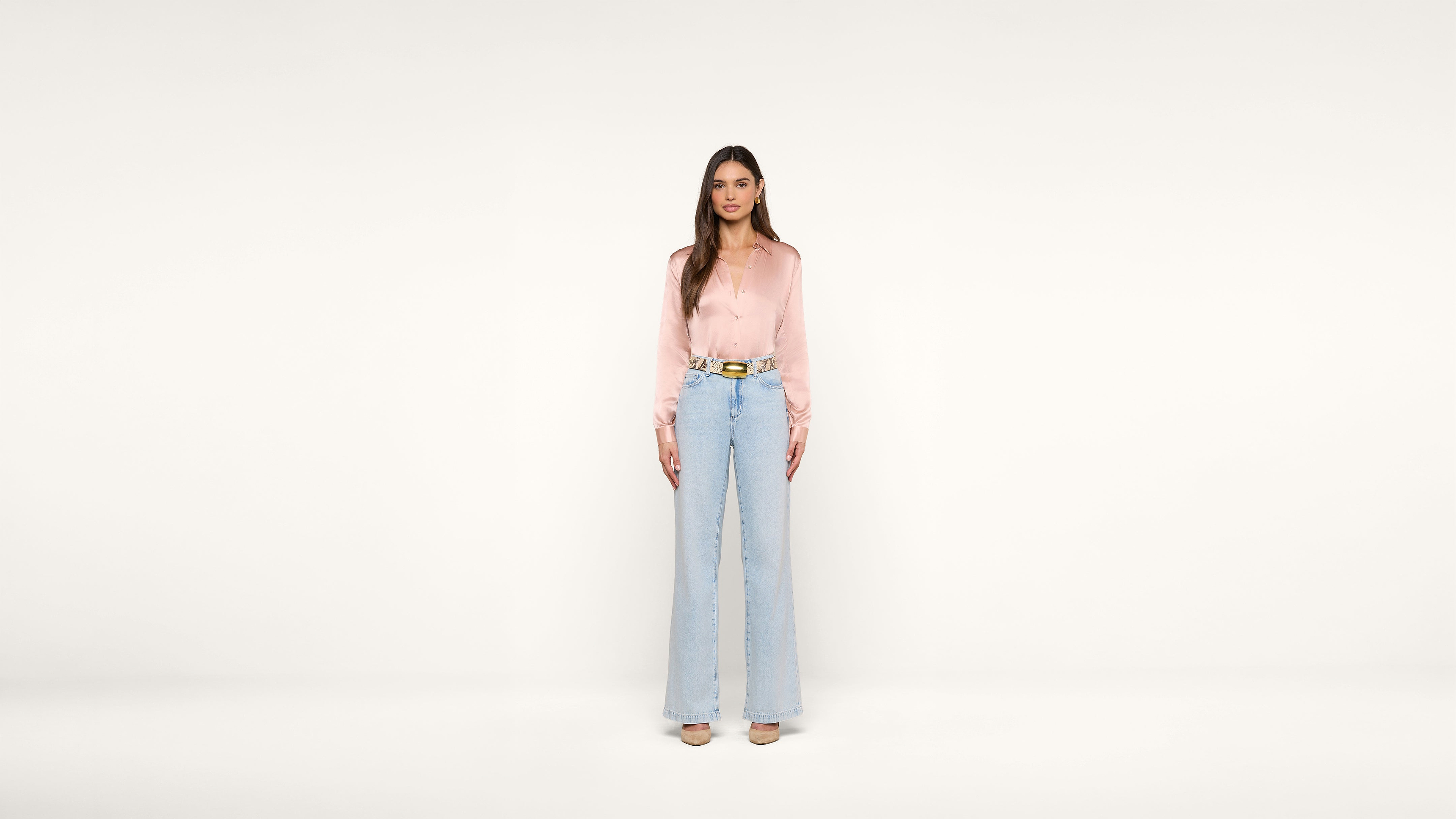 A woman stands facing forward against a plain white background, wearing a light pink blouse, light blue high-waisted wide-leg jeans, and beige shoes.