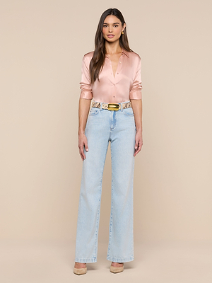 A woman stands facing forward, wearing a shiny pink Tyler Silk Blouse, light blue high-waisted wide-leg jeans, beige heels, and a patterned belt with a gold buckle against a beige background.