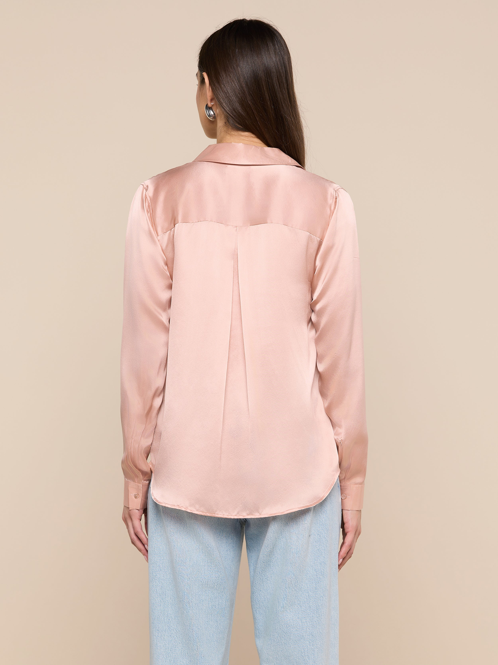 A woman with long brown hair is seen from behind, wearing the Tyler Silk Blouse in light pink with long sleeves and light blue jeans, standing against a beige background.