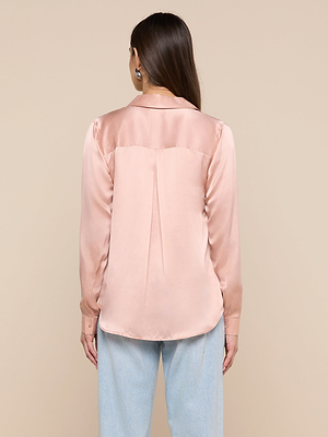 A woman with long brown hair is seen from behind, wearing the Tyler Silk Blouse in light pink with long sleeves and light blue jeans, standing against a beige background.