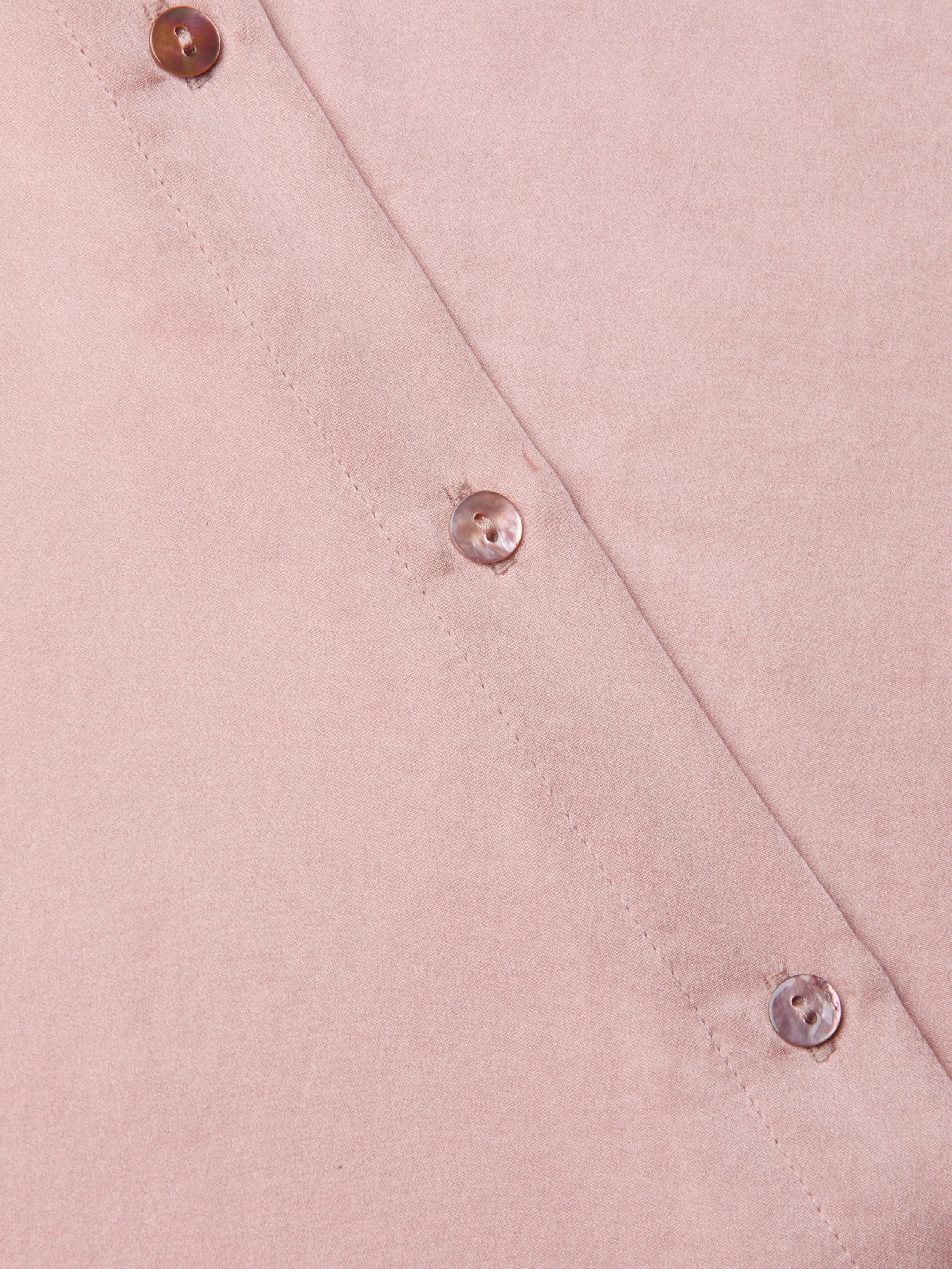 Close-up of the Tyler Silk Blouse in light pink, featuring three shiny round buttons arranged diagonally, showcasing its smooth silk texture and precise stitching along the edge.