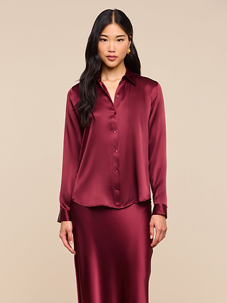 A woman with long black hair wears the Tyler Silk Blouse in shiny, long-sleeve merlot red silk charmeuse, paired with a matching skirt, standing against a plain beige background.