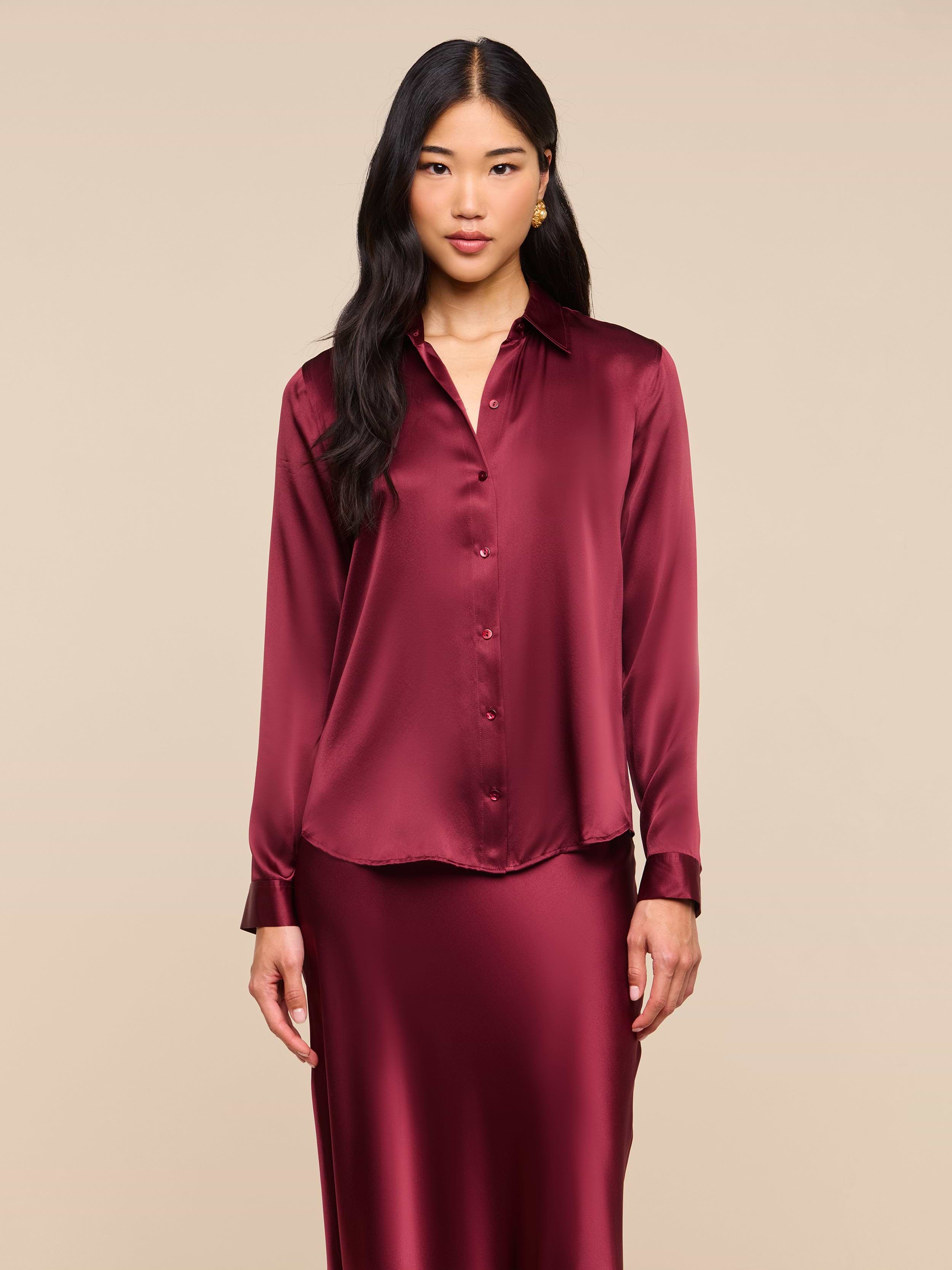A woman wearing the Tyler Silk Blouse—a long-sleeved, button-up silk charmeuse top in merlot red—and a matching skirt, stands against a plain light background, looking at the camera.