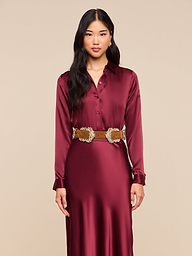 A woman in the Tyler Silk Blouse, a long-sleeved merlot red silk charmeuse top paired with a gold belt, stands facing forward against a plain light background.