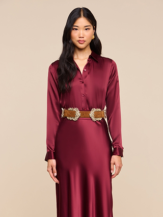 Woman in the Tyler Silk Blouse in merlot red, styled with a brown belt featuring ornate silver buckles, standing against a beige background.