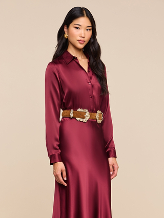 A woman wearing the Tyler Silk Blouse in merlot red, styled with a tan belt featuring large silver decorative buckles, stands against a plain beige background.