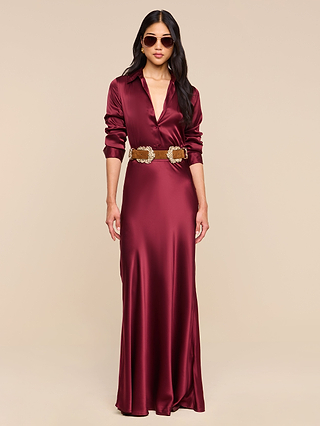 A woman wearing the Tyler Silk Blouse in merlot red, styled with a tan belt and sunglasses, with long dark hair, standing against a beige background.