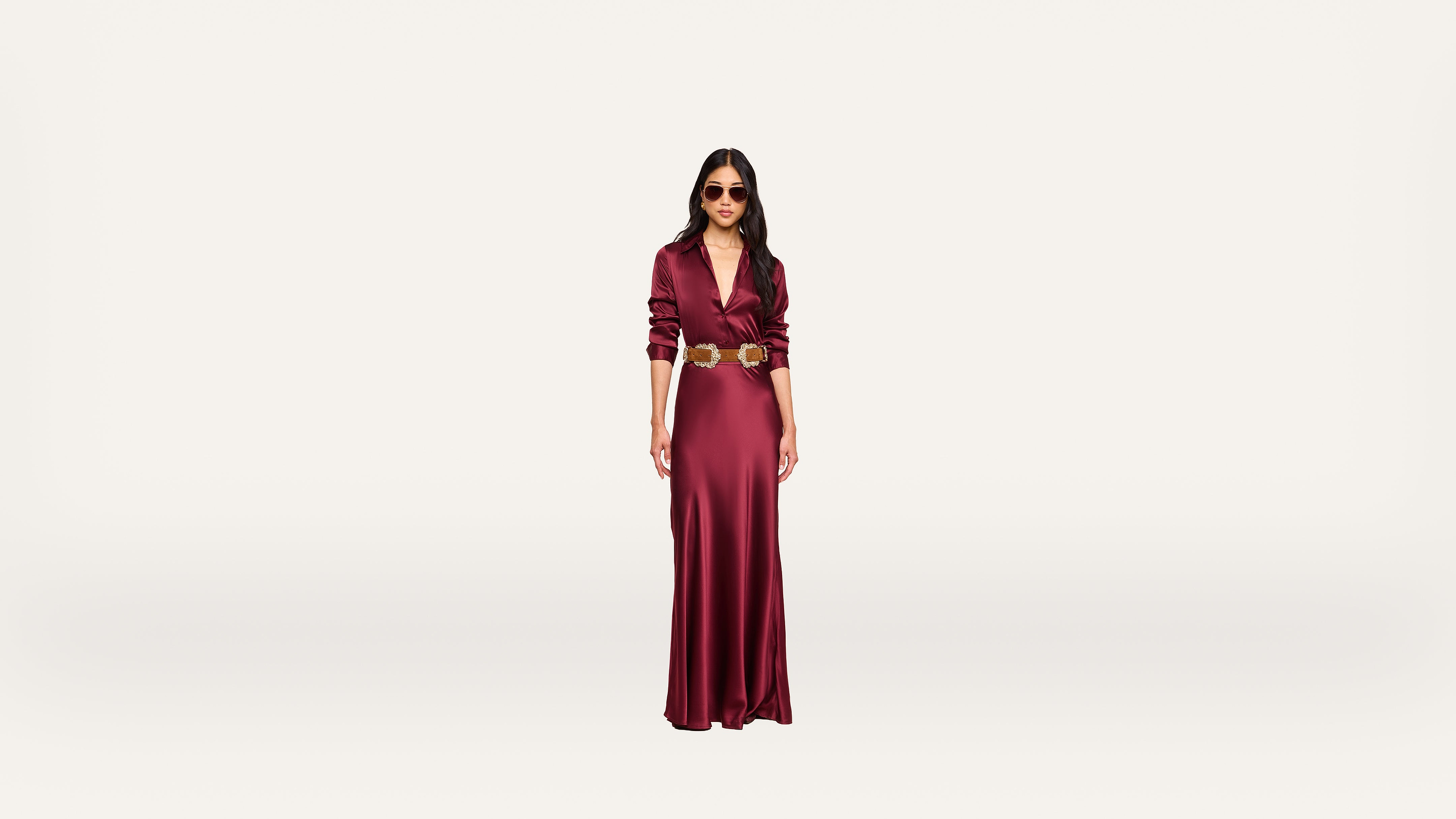 A woman wearing a long, deep red dress with a gold belt and sunglasses stands against a plain white background.