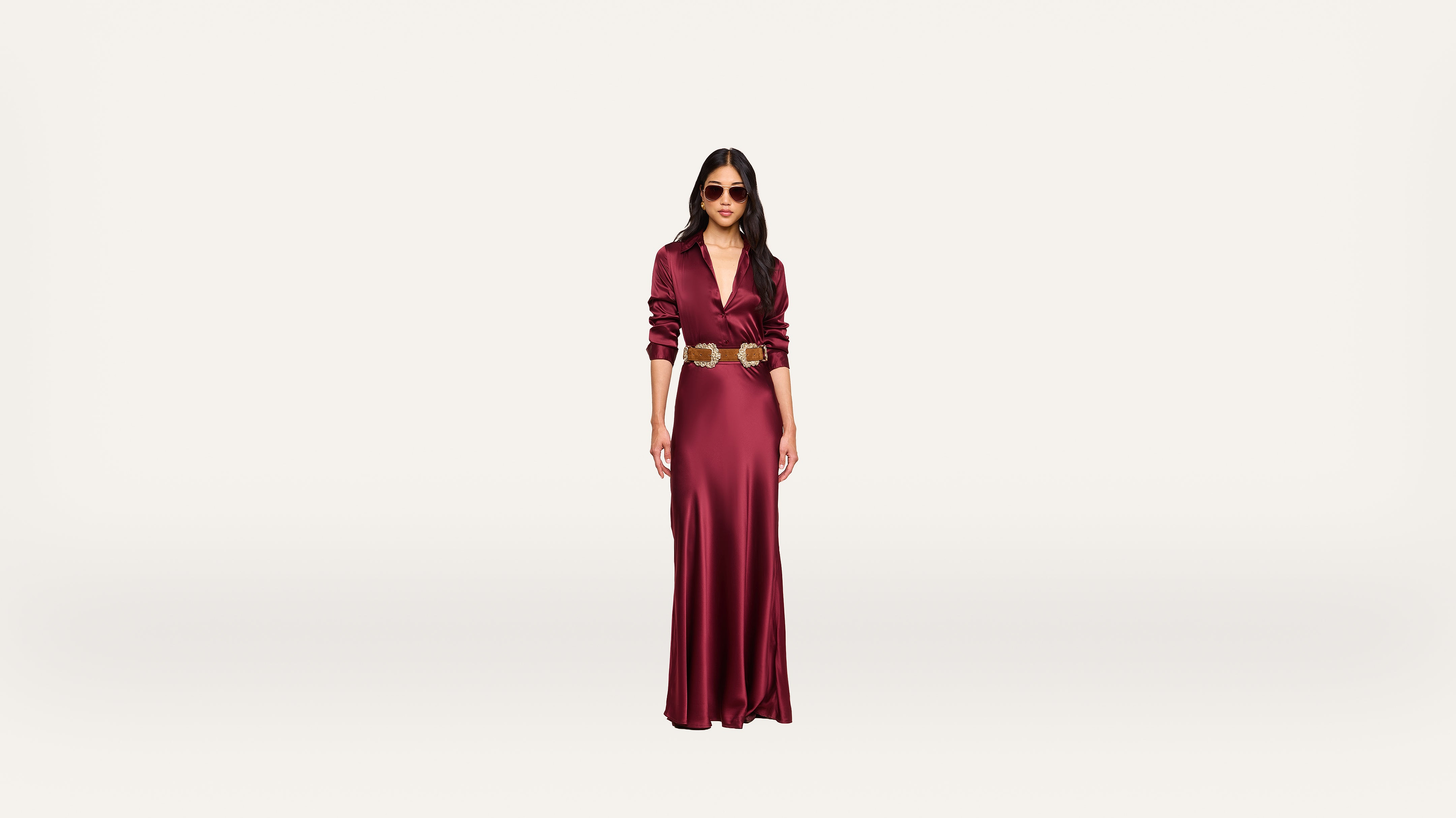 A woman wearing a long, deep red dress with a gold belt and sunglasses stands against a plain white background.